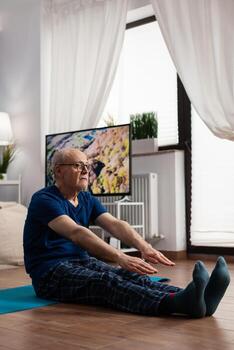 mayor hombre extensión piernas músculo mientras sentado en yoga estera en vivo habitación durante pilates ejercicio. pensionista formación cuerpo flexibilidad adelgazar peso hacer ejercicio gimnástico ejercicio foto