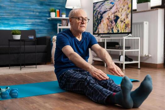 Jubilación mayor hombre sentado en yoga estera extensión piernas músculos durante cuerpo formación trabajando a flexibilidad. pensionista en ropa de deporte adelgazar peso durante músculo resistencia en vivo habitación foto