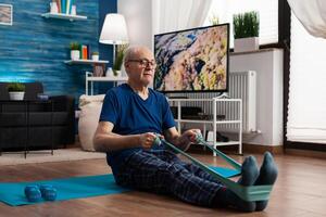 Retirement senior man sitting on yoga mat stretching legs muscles using resistance elastic band training body flexibility. Pensioner in sportswear slimming weight during muscle training in living room photo