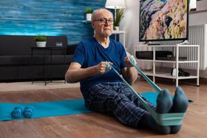 Focused pensioner sitting on yoga mat stretching legs muscle using fitness elastic band during aerobics workout exercising muscles resistance. Senior man doing body exercise slimming weight photo