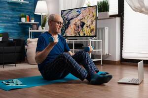 Retired senior man sitting on yoga mat stretching abdominals muscles practicing fitness workout using dumbbells. Pensioner working at body warming up during wellness routine watching pilates photo