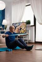 Retirement senior man sitting on yoga mat with leg in crossed position stretching arms muscles using stretch elastic band during sport routine in living room. Pensioner exercising bodyresistance photo
