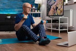 Senior man in sportwear warming up body muscles practicing wellness exercise using workout dumbbells. Pensioner sitting on yoga mat with crossed legs during aerobics routine looking at gym photo
