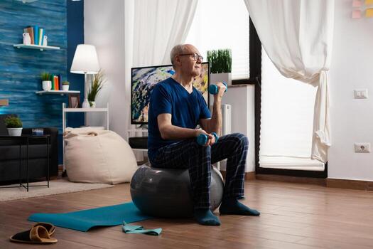 Jubilación mayor hombre sentado en suizo pelota hacer ejercicio brazos músculos haciendo aptitud ejercicios utilizando rutina de ejercicio mancuernas enfocado pensionista formación cuerpo fuerza resistencia en vivo habitación foto