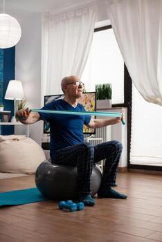 alegre retirado mayor hombre hacer ejercicio brazo músculos utilizando resistencia elástico banda practicando aerobio ejercicio. pensionista sentado en suizo pelota en vivo habitación trabajando a cuerpo cuidado de la salud foto