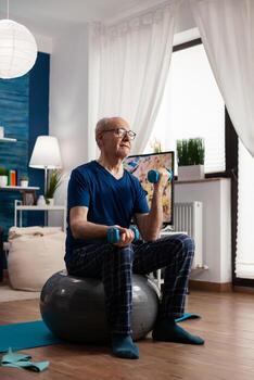 mayor hombre participación pesas haciendo brazos ejercicio estirar cuerpo músculos formación muscular resistencia. Jubilación caucásico masculino sentado en suizo pelota en vivo habitación haciendo aptitud rutina de ejercicio foto
