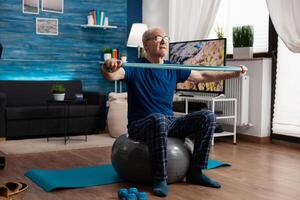Cheerful retired senior man exercising arm muscles using resistance elastic band practicing aerobic exercise. Pensioner sitting on swiss ball in living room working at body healthcare photo