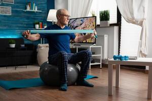 Invalid pensioner practicing active arm exercises using resistance elastic band watching online fitness workout on laptop. Pensioner working body healthcare sitting on swiss ball in living room photo