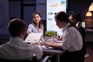 Executive manager woman explaining management statistics working at company strategy overtime in office meeting room. Diverse multi-ethnic businesspeople teamwork brainstorming ideas in evening. photo