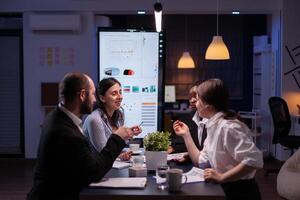 Diverse multi-ethnic businesspeople analyzing management strategy working overtime in company office meeting room late at night. Overworked leader overworking marketing strategy in evening photo