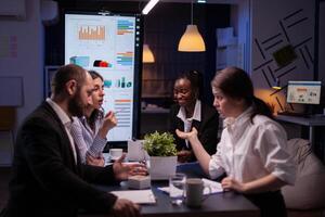Diverse multi-ethnic businesspeople analyzing management strategy working overtime in company office meeting room late at night. Overworked leader overworking marketing strategy in evening photo