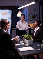 Executive manager woman explaining management statistics working at company strategy overtime in office meeting room. Diverse multi-ethnic businesspeople teamwork brainstorming ideas in evening. photo