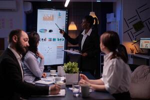 Workaholic african american leader standing in front of presentation monitor explaining marketing project solution late at night in company meeting room. Diverse teamwork working at business ideas. photo