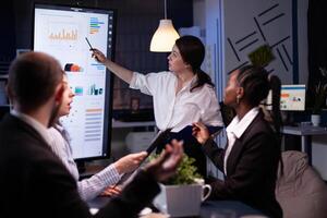 Multi ethnic businesspeople brainstorming company ideas checking graphs statistics using tv standing at conference table. Diverse teamwork working at management presentation in meeting room photo