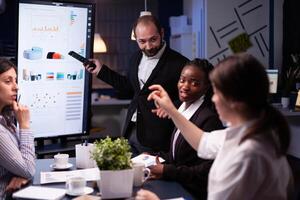 Entrepreneur man brainstorming management strategy working hard in meeting office room late at night. Diverse multi-ethnic business team looking at financial company presentation on monitor. photo