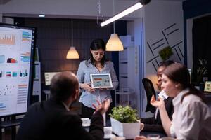 Overworked workaholic entrepreneur woman shwoing marketing graphs using tablet overworking at company solution late at night in meeting room. Diverse multi-ethnic teamwork solving financial strategy photo