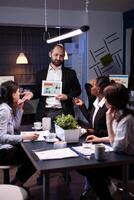 Business entrepreneur man presenting company statistics using tablet for financial presentation. Diverse multi-ethnic businesspeople brainstorming strategy working in office meeting room late at night photo