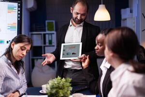 Focused workaholic entrepreneur man working overtime presenting company statistics using tablet. Diverse multi-ethnic businesspeople overworking in office meeting room late at night. photo