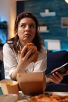 Caucasian female holding tastu burgery in hands browsing on social media using smartphone relaxing on couch during junck-food night. Woman enjoying takeaway food home delivery photo