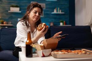 Caucasian female holding tastu burgery in hands browsing on social media using smartphone relaxing on couch during junck-food night. Woman enjoying takeaway food home delivery photo