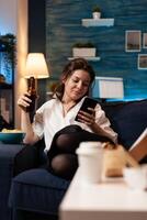 Caucasian female relaxing on couch browsing on social media using smarhphone while drinking beer during relaxing night at home. Woman enjoying takeaway food home delivered. Junk-food order photo