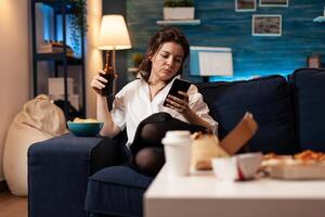 Caucasian female relaxing on couch browsing on social media using smarhphone while drinking beer during relaxing night at home. Woman enjoying takeaway food home delivered. Junk-food order photo