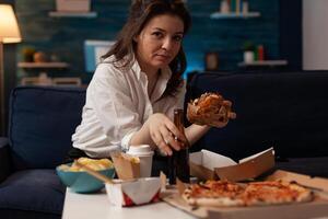 Woman holding beer bottle eating tasty takeaway hamburger while relaxing on couch in front of tv table with fast food menu. Hungry person enjoying junk food sitting on sofa in the evening. photo