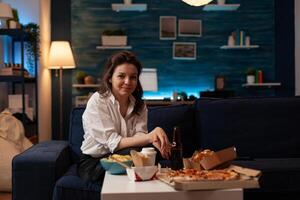 Portrait of smiling woman relaxing on couch after office work enjoying fast food dinner in the evening. Fast-food takeaway menu. Junk food order on coffee table. photo