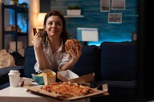 Woman eating tasty takeaway fast food hamburger while enjoying a cold beer in front of television with comedy show in home living room. Smiling person having hot pizza and takeout delivery menu. photo