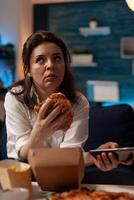 Portrait of woman holding tasty burger takeaway dinner browsing on smartphone looking bored sitting on sofa in home living room. Person having home deliverd fast food menu while browsing social media. photo