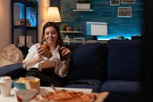 Happy woman eating tasty takeaway hamburger holding beer bottle relaxing on couch watching television family sitcom. Smiling person having takeout burger and fast food delivery menu. photo