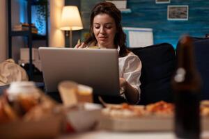 Woman browsing social media on laptop while having fast food snack sitting on couch in living room. Office worker with personal computer eating chips in front of table with home delivery menu. photo