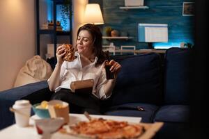 Smiling woman sitting on couch looking at tasty takeaway burger holding beer bottle in front of table with pizza and takeaway junk food menu. Happy person eating tasty burger and delivery dinner. photo