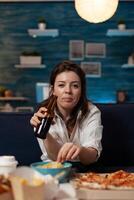 Portrait of woman looking at camera holding beer bottle and eating potato chips from bowl at table with large pizza and takeaway dinner. Office worker on couch relaxing after takeout menu. photo