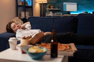Person sleeping on sofa after drinking beer and eating home delivery pizza in front of television in living room. Tired woman falling asleep couch after large fast food takeout meal. photo