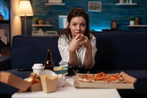 Person sitting on couch in living room having a slice of delivery pizza while watching television sitcom at table with fast food takeout menu. Woman after office work eating takeaway tv dinner. photo