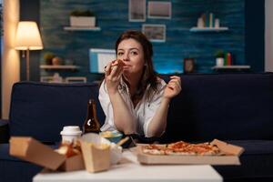 Woman after office work taking a bite out of slice of hot delivery pizza while watching television at table with fast food takeway menu. Person sitting on couch in living room eating takeout dinner. photo