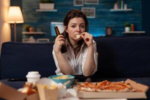 Sad woman drinking craft beer and eating potato chips from bowl at table with large pizza and takeout dinner in home living room. Tired office worker on couch relaxing after takeaway menu. photo