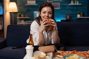 Smiling woman holding tasty burger in hands looking at television comedy sitcom standing on couch in front of table with takeaway fast food menu. Happy person watching tv show having takeout dinner. photo