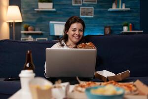 Portrait of smiling woman watching online television sitcom on laptop eating a home delivery burger for dinner. Happy person holding portable computer eating takeaway fast food menu. photo