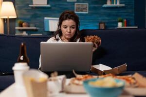 Influencer checking social media on laptop eating a tasty takeout burger and fast food delivery menu in living room. Content creator watching online series while having takeaway hamburger. photo
