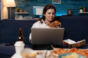 Programmer relaxing on couch holding laptop eating a tasty takeaway burger and fast food delivery menu in living room. Coder after work watching online series while having a hamburger. photo