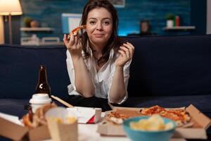 Portrait of smiling woman after office work eating a slice of hot delivery pizza while watching television at table with fast food takeaway menu. Happy person sitting on couch eating takeout tv dinner. photo