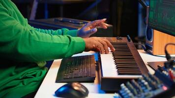 African american audio technician using piano synthesizer keys to add sound effects in post production, editing tunes with digital software and mixing console. Male artist in home studio. Camera A. photo