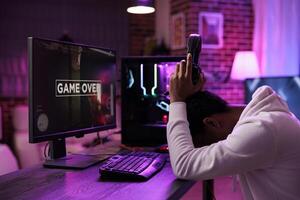 Sad gamer upset after losing online multiplayer action videogame championship, being defeated by rivals. Man removing headphones in frustration after seeing game over screen on gaming pc display photo
