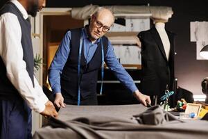 Experienced elderly master suitmaker preparing new fabric sheet for upcoming sartorial collection manufacturing. Senior tailor and apprentice ready to measure, cut, sew and stich textile material photo
