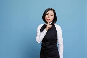 Asian waitress dressed in restaurant uniform showing silent gesture and looking at camera with serious face expression. Woman receptionist making tsss sign while holding finger on lips portrait photo