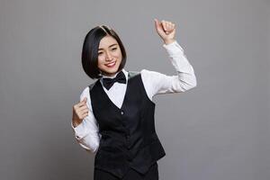 Restaurant cheerful smiling asian waitress dancing, raising hands with positive facial expression. Carefree relaxed attractive woman receptionist moving to music rhythm in studio photo