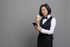 Carefree asian woman in waitress uniform drinking coffee and using social media app on smartphone. Catering service smiling worker holding tea to go paper cup and communicating online on mobile phone photo