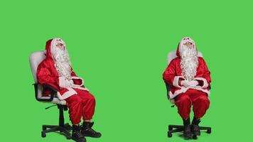 Saint nick embodiment sitting on chair, posing and waiting for something dressed with red suit and hat with white fake beard. Santa iconic character looking around the studio, full body backdrop. photo